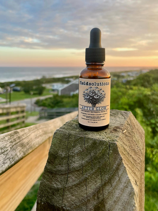 After Glow chamomile and jojoba face serum in an amber glass dropper bottle on a weathered wooden post above a coastal view at sunset